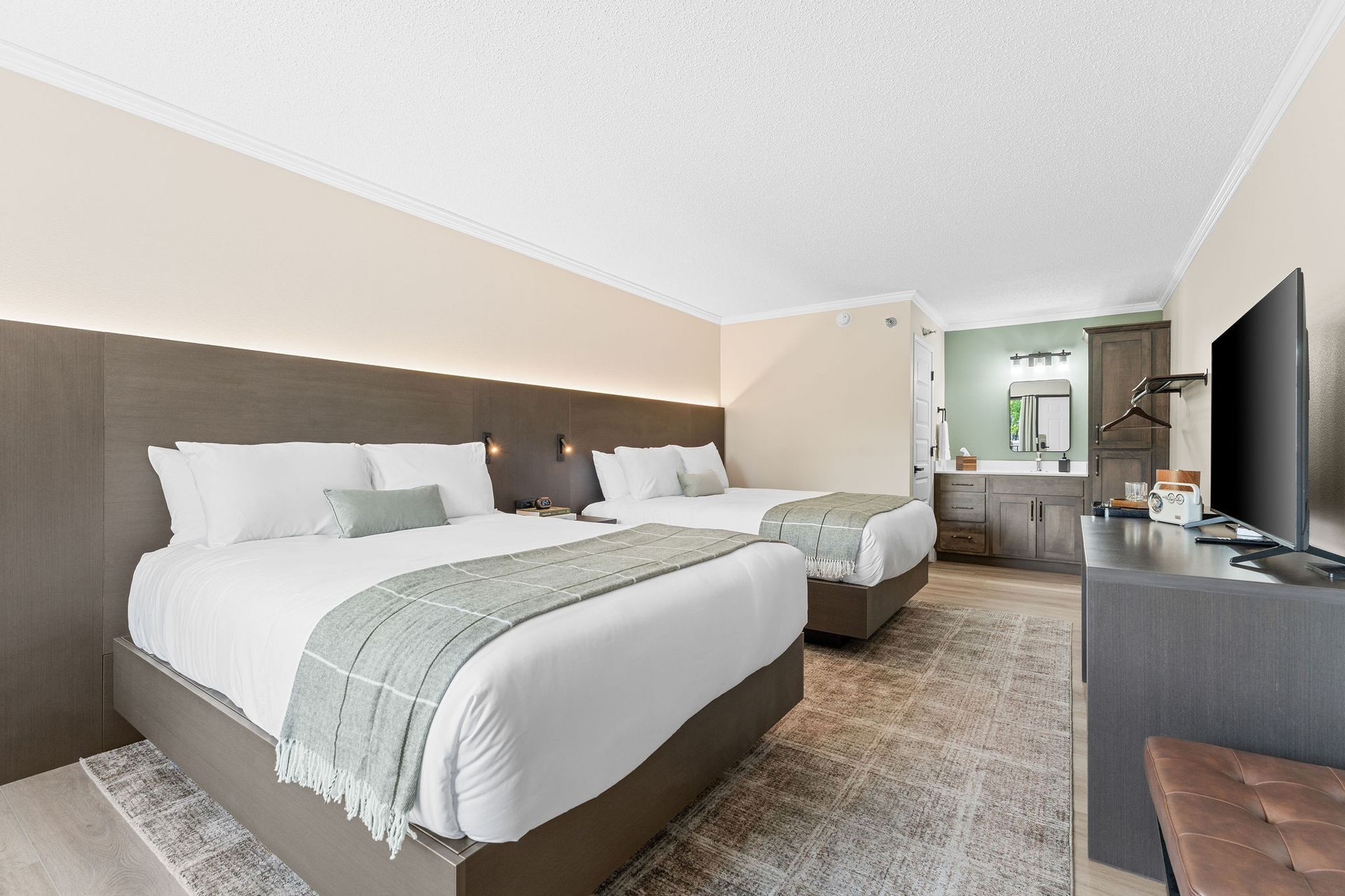 A hotel room with two queen beds featuring white bedding, gray accents, and a television, near an open bathroom vanity.