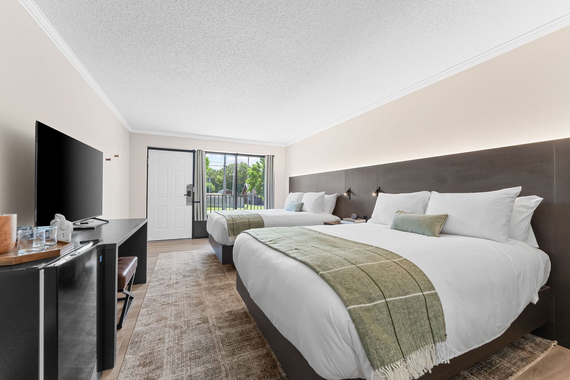 A hotel room with two white beds, a television on a dark desk, and a bright window with green trees visible outside.