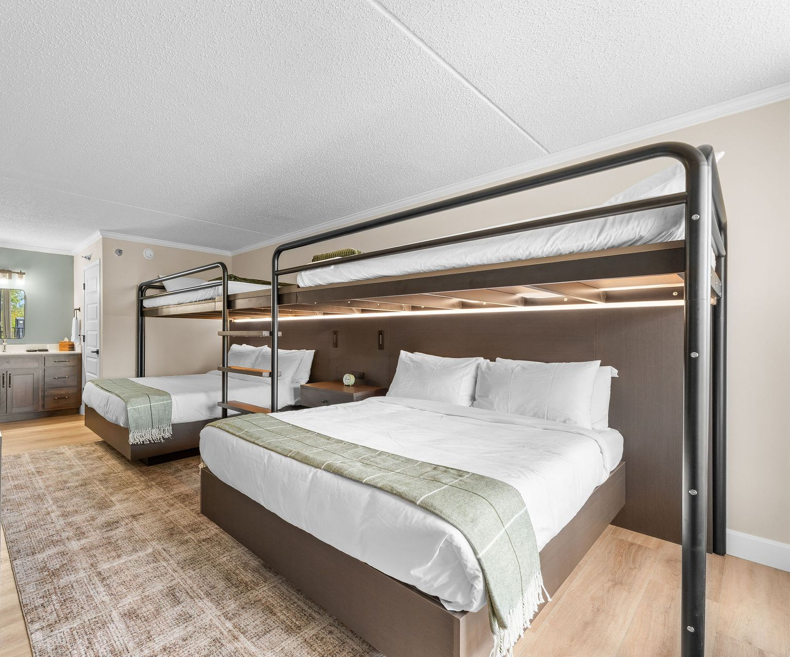 A hotel room featuring two queen-size bunk beds with white bedding, light wood floors, and a textured area rug.
