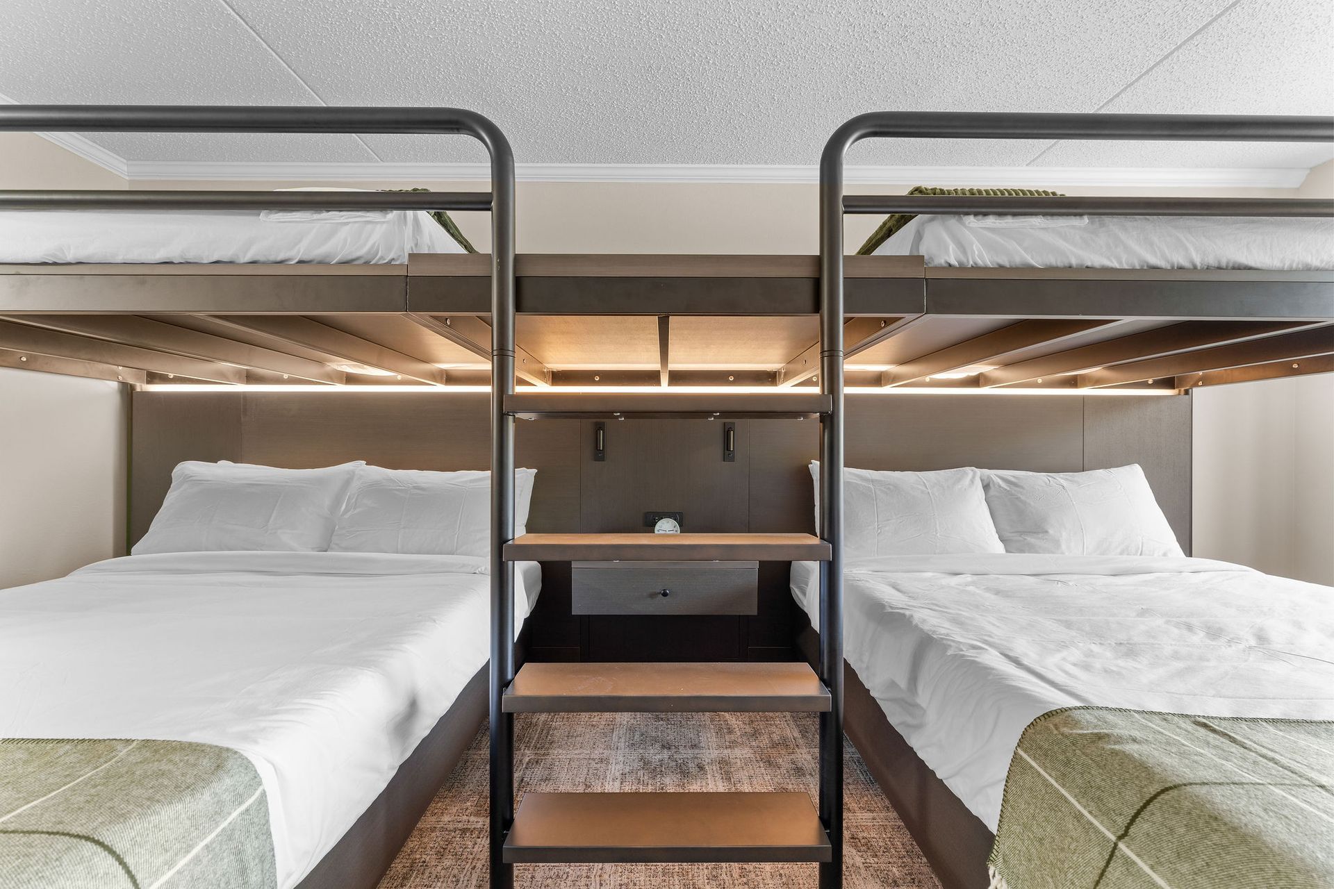 Two queen beds in a hotel room, each with a built-in metal bunk bed above, separated by a central ladder and shelving unit.
