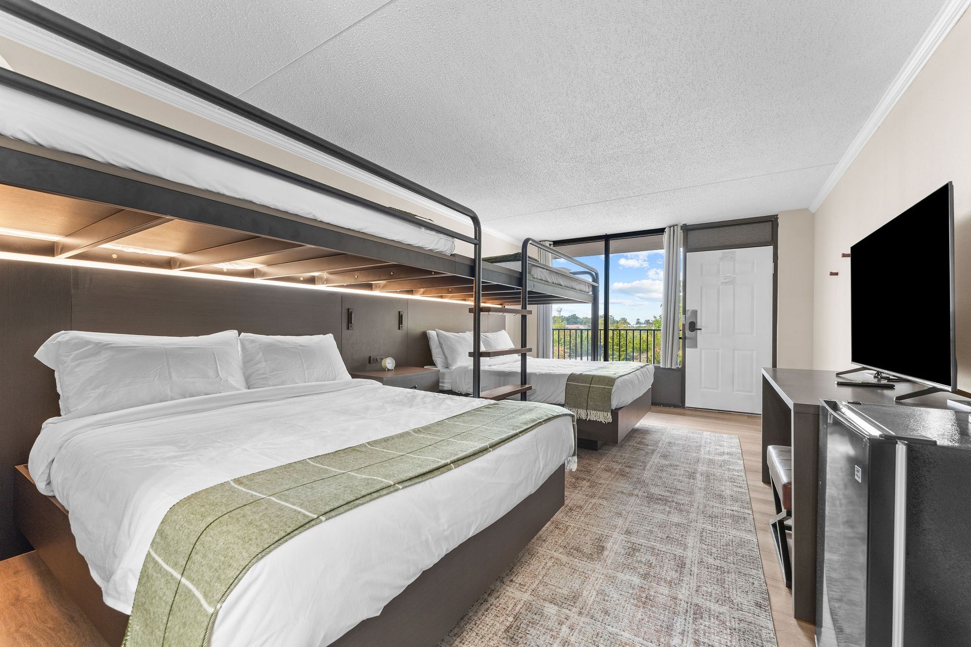A hotel room featuring a bunk bed setup above a queen bed, a second bed in the back, a TV, and a mini-fridge.