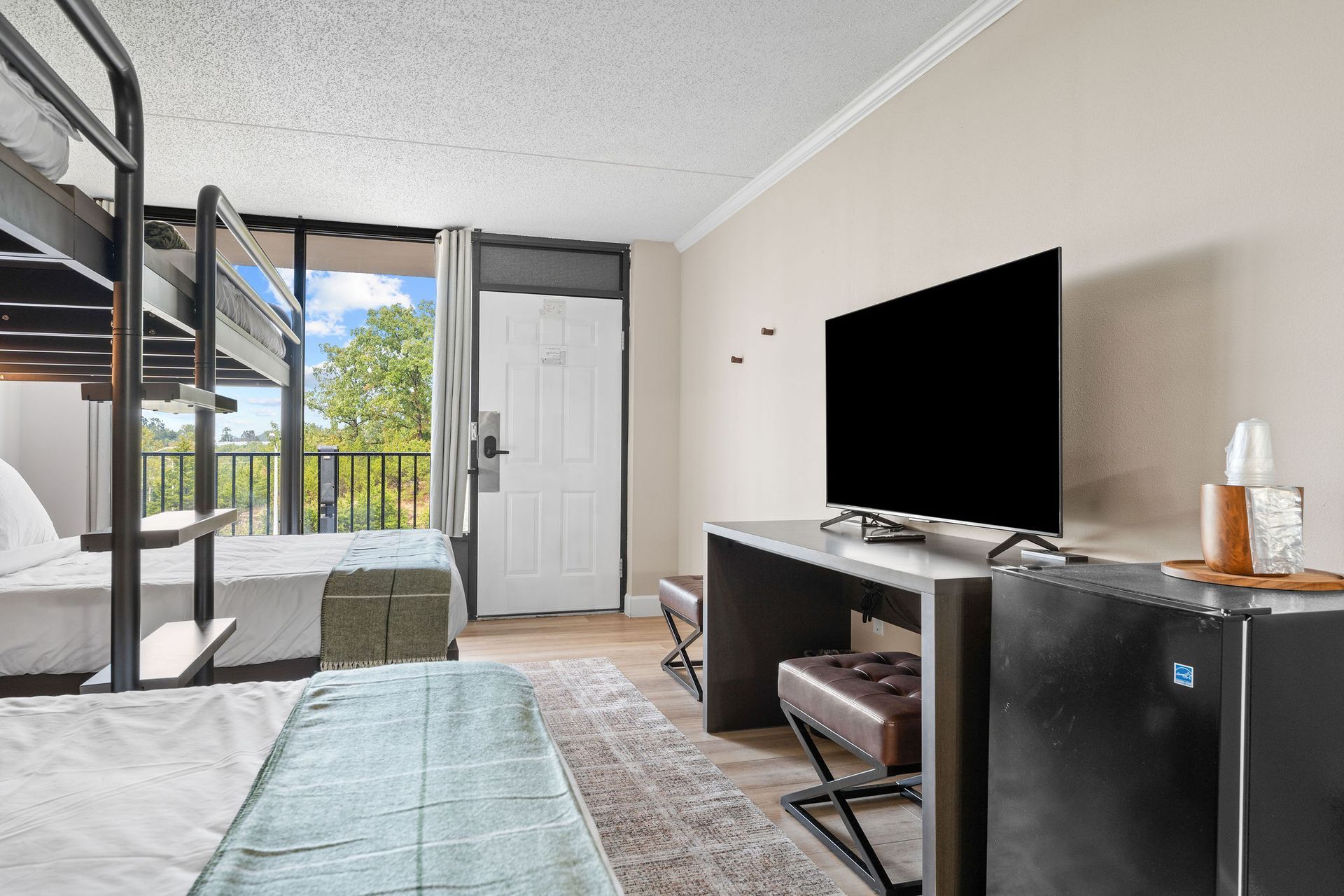 A hotel room featuring a bunk bed, a wall-mounted flat-screen television above a desk, a mini-fridge, and a balcony door.