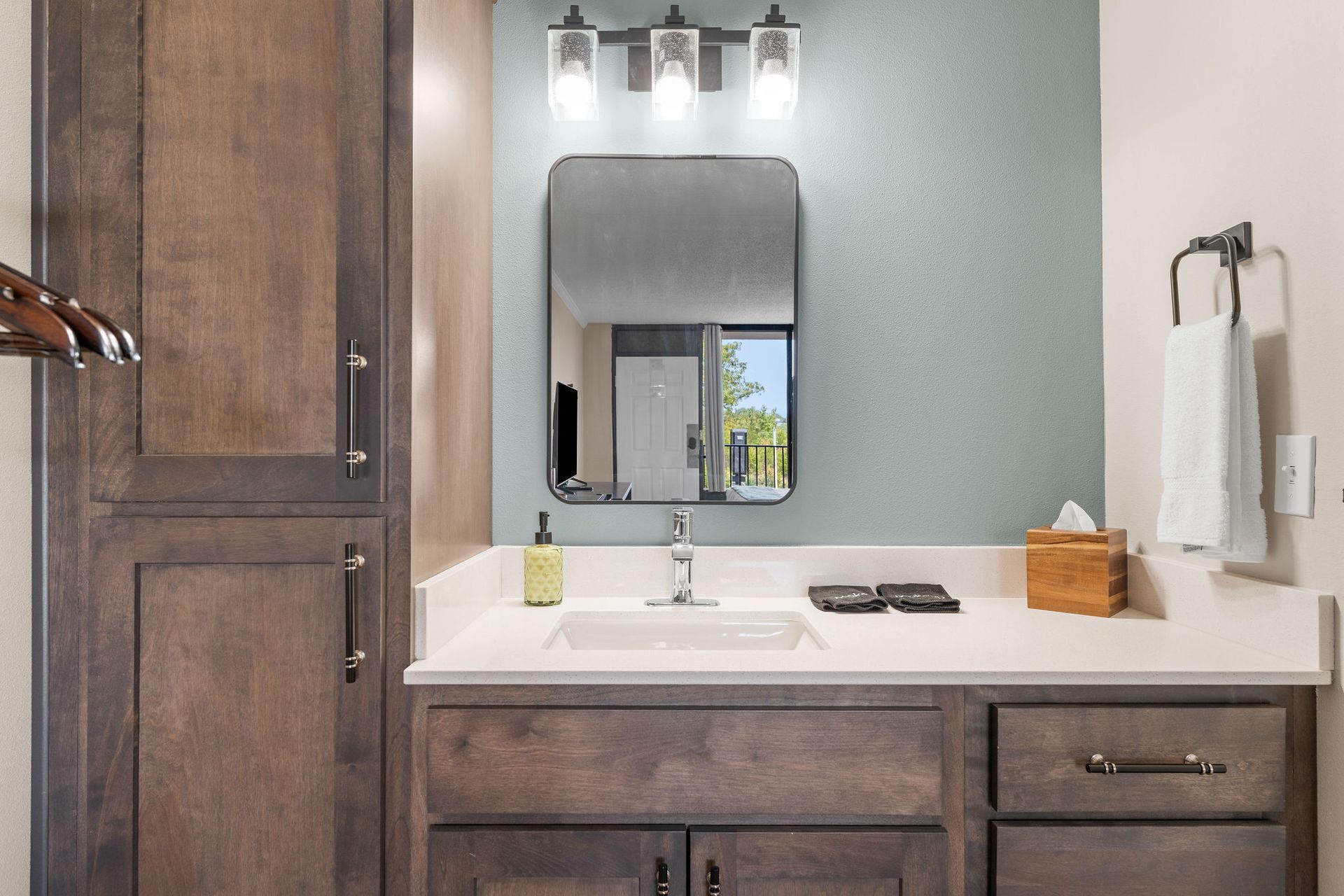 A hotel bathroom vanity featuring a light stone countertop, wood cabinetry, a rectangular mirror, and sage green wall.