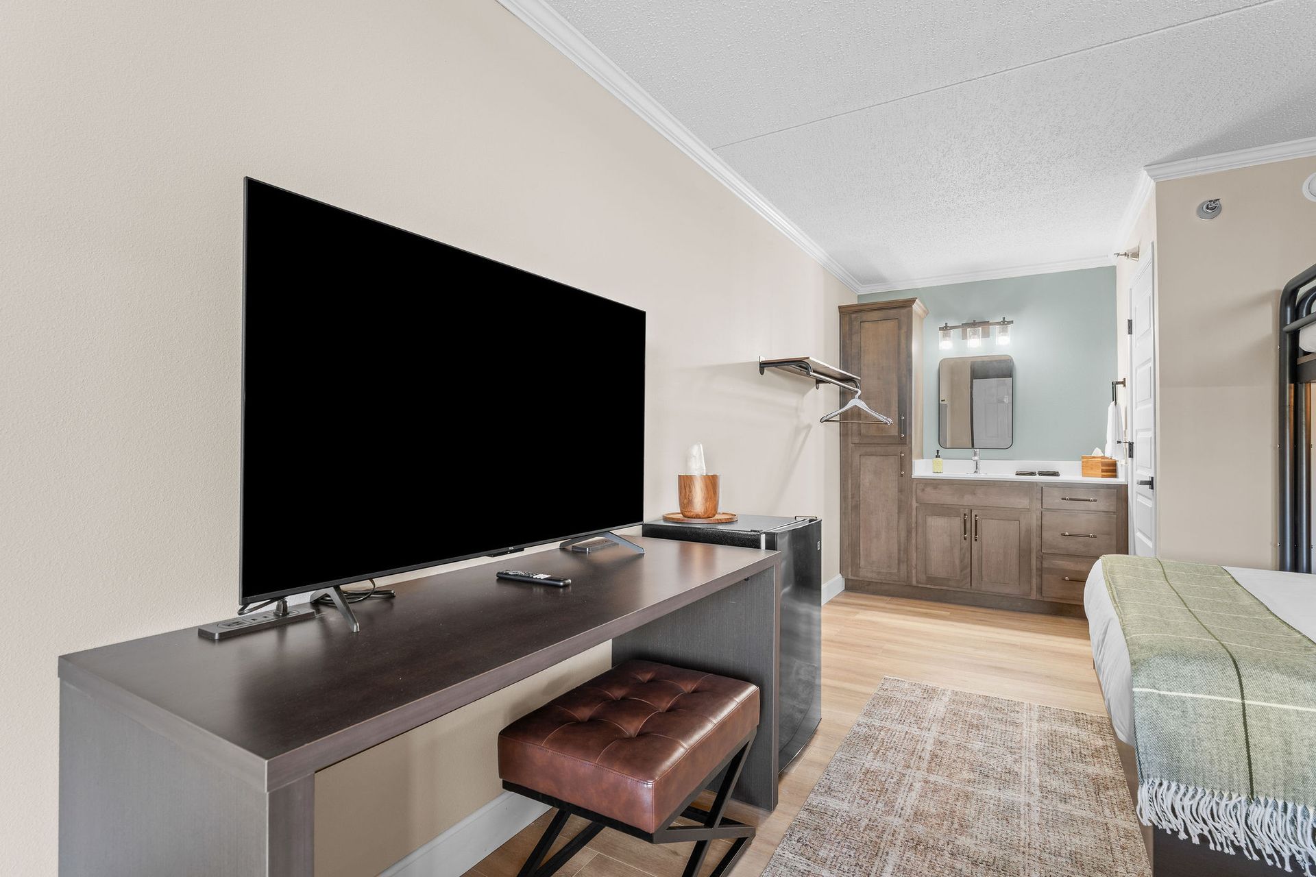 A bedroom featuring a flat-screen TV on a desk, a brown leather stool, a vanity area, and a bed corner.