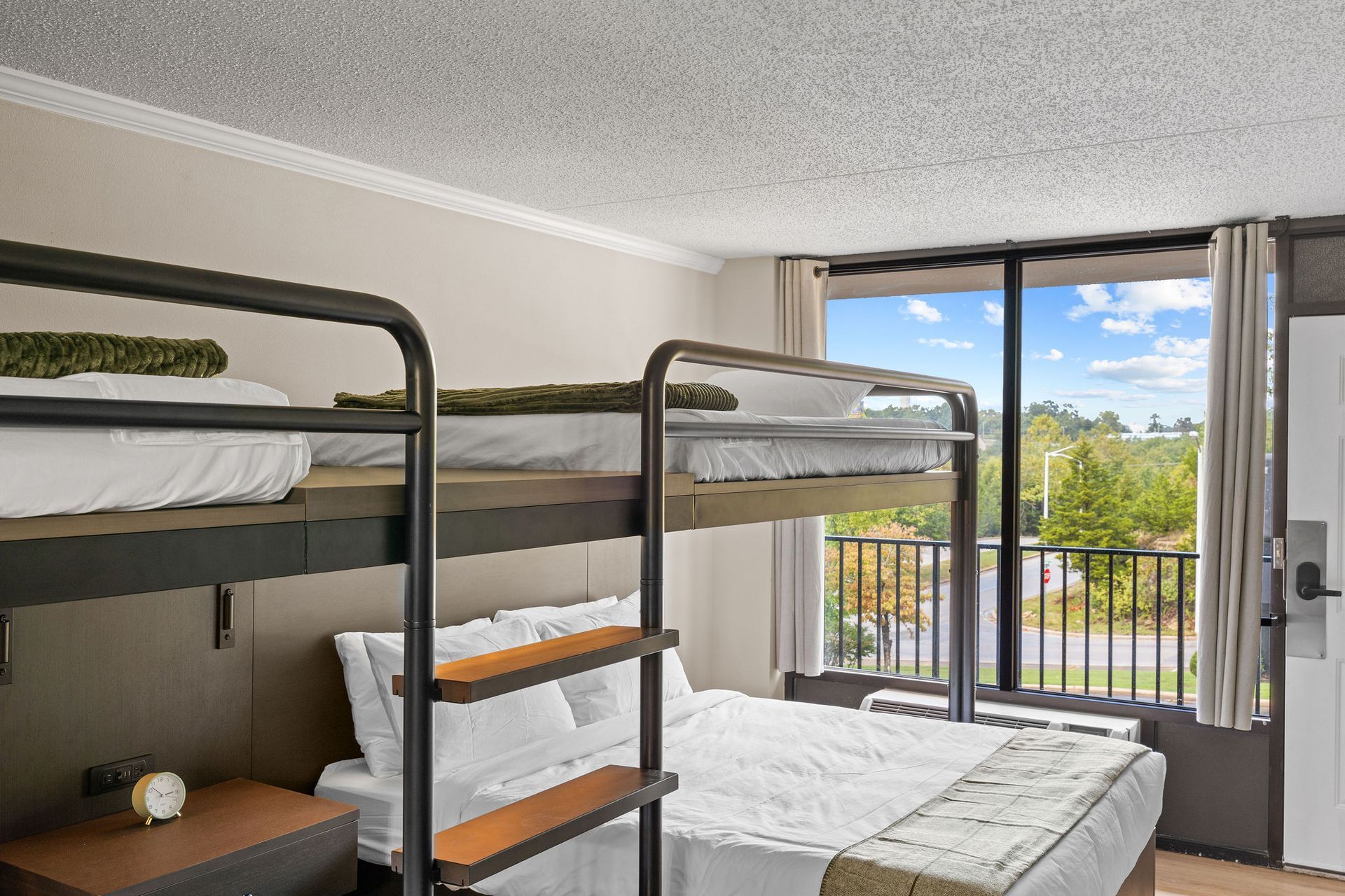 A modern hotel room featuring a bunk bed over a queen bed, next to a large window overlooking trees and a parking lot.