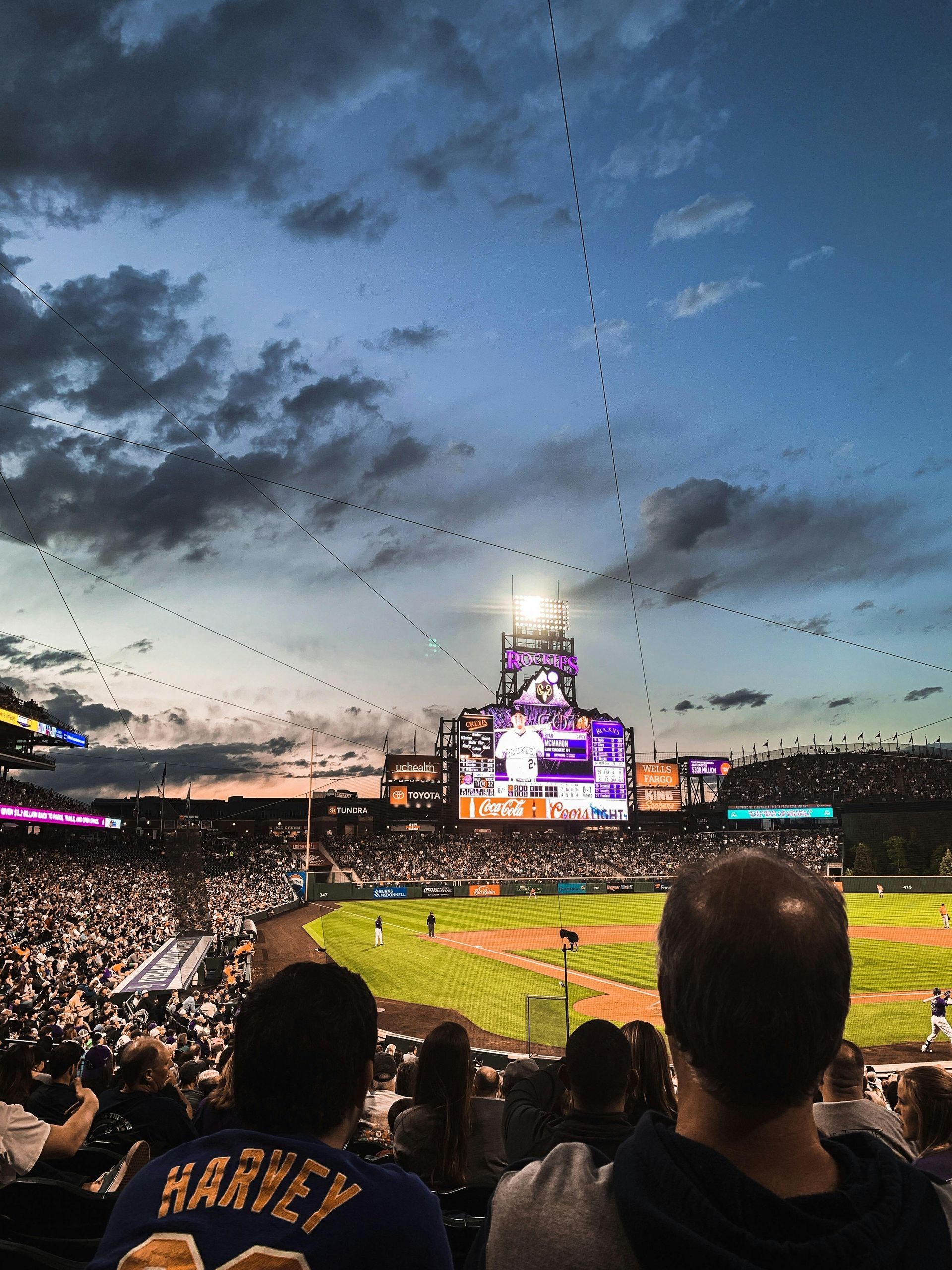View from the stands at a baseball game at dusk, featuring a large illuminated scoreboard and a string of lights.