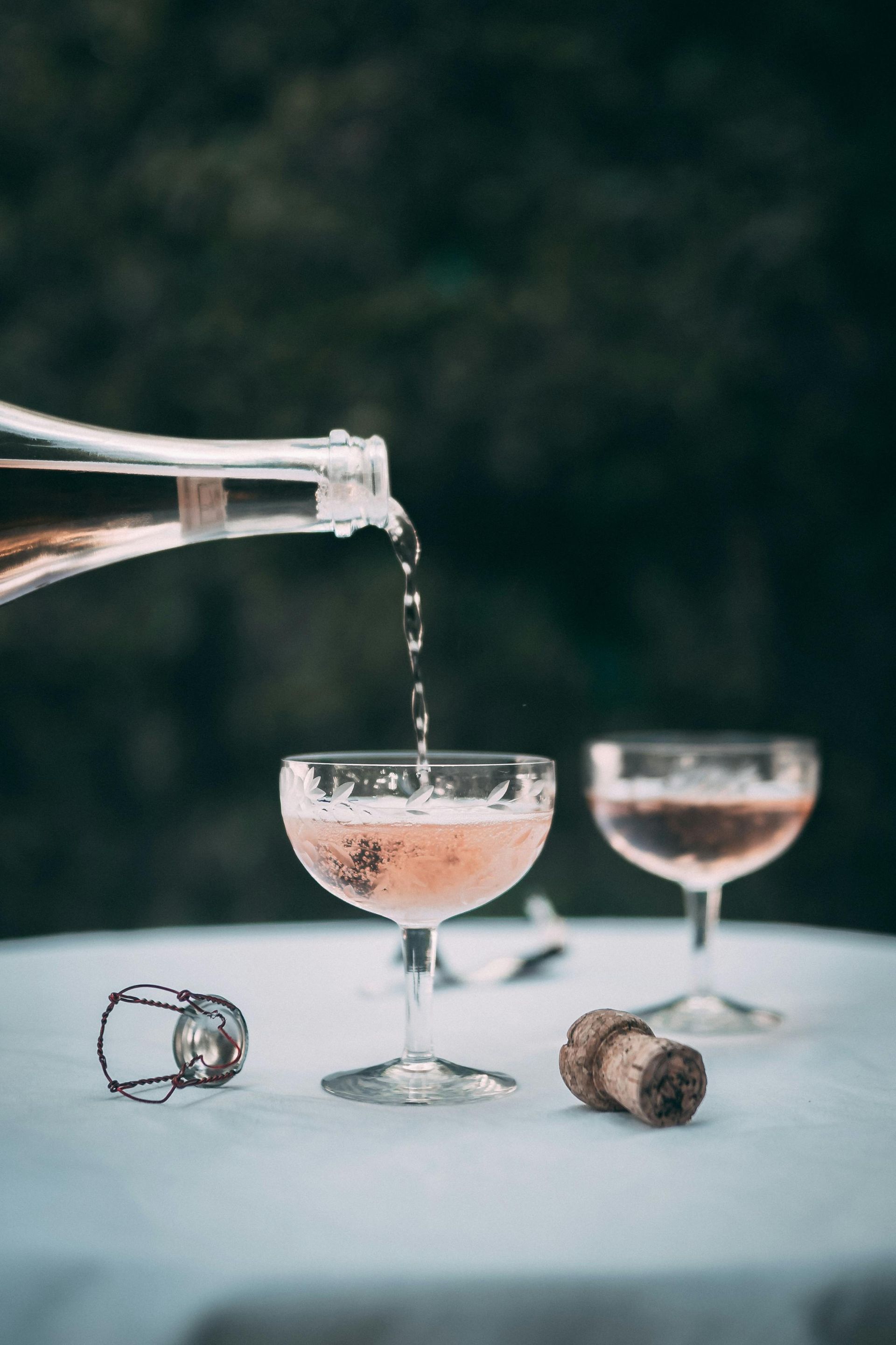 A bottle pours sparkling rosé into a glass coupe on a white tablecloth, with another glass and a cork nearby.