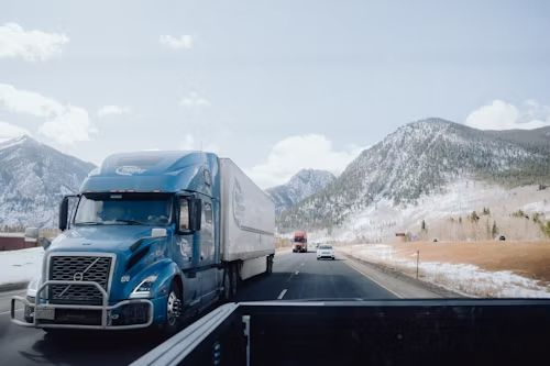 Blue semi-truck driving on a highway with snowy mountains in the background. Sunny day.