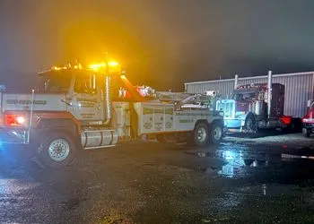 Tow truck towing a semi-truck at night, flashing amber lights, on wet asphalt.