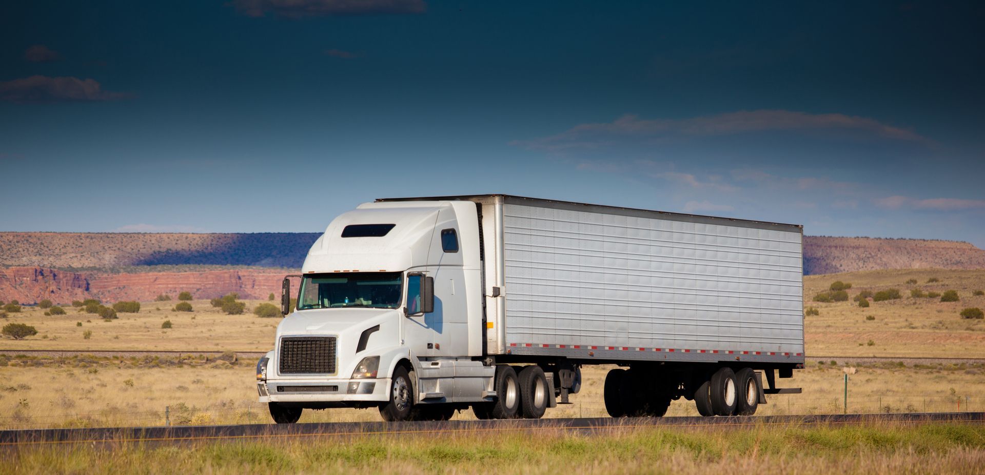 A white semi truck is driving down a road in the desert.
