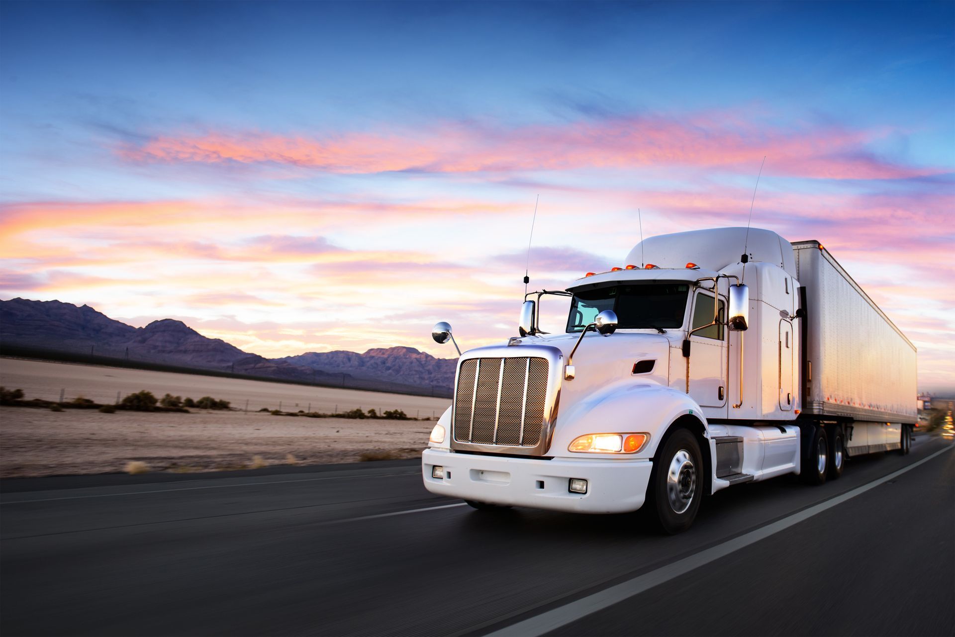 A white semi truck is driving down a highway at sunset.