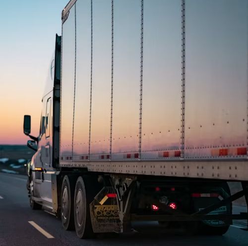 Semi-truck trailer driving on a road at dusk, with sunset reflections on its white side.