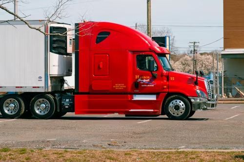Red semi-truck with white trailer parked in a lot. The truck has the company logo on its side.