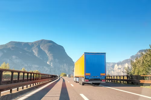 A semi-truck with a yellow trailer and blue back drives on a highway surrounded by mountains under a clear blue sky.