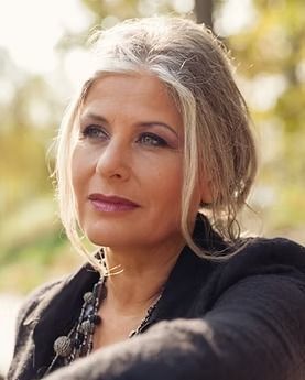 Portrait of a woman outdoors with gray hair, wearing a black jacket and necklace, looking calmly to the side