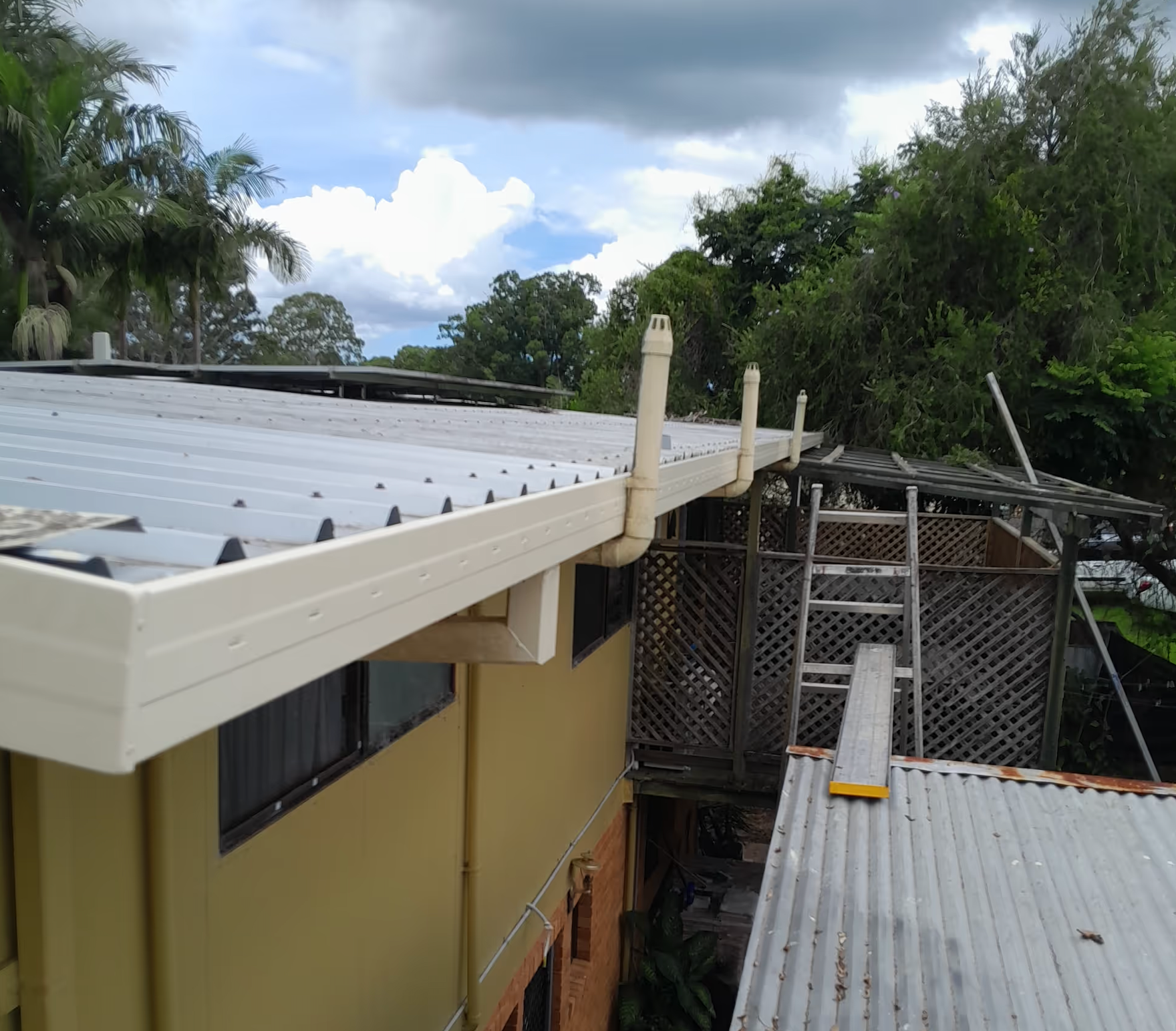Roofing over a narrow alley beside a yellow house, with corrugated metal sheets and a ladder under cloudy skies