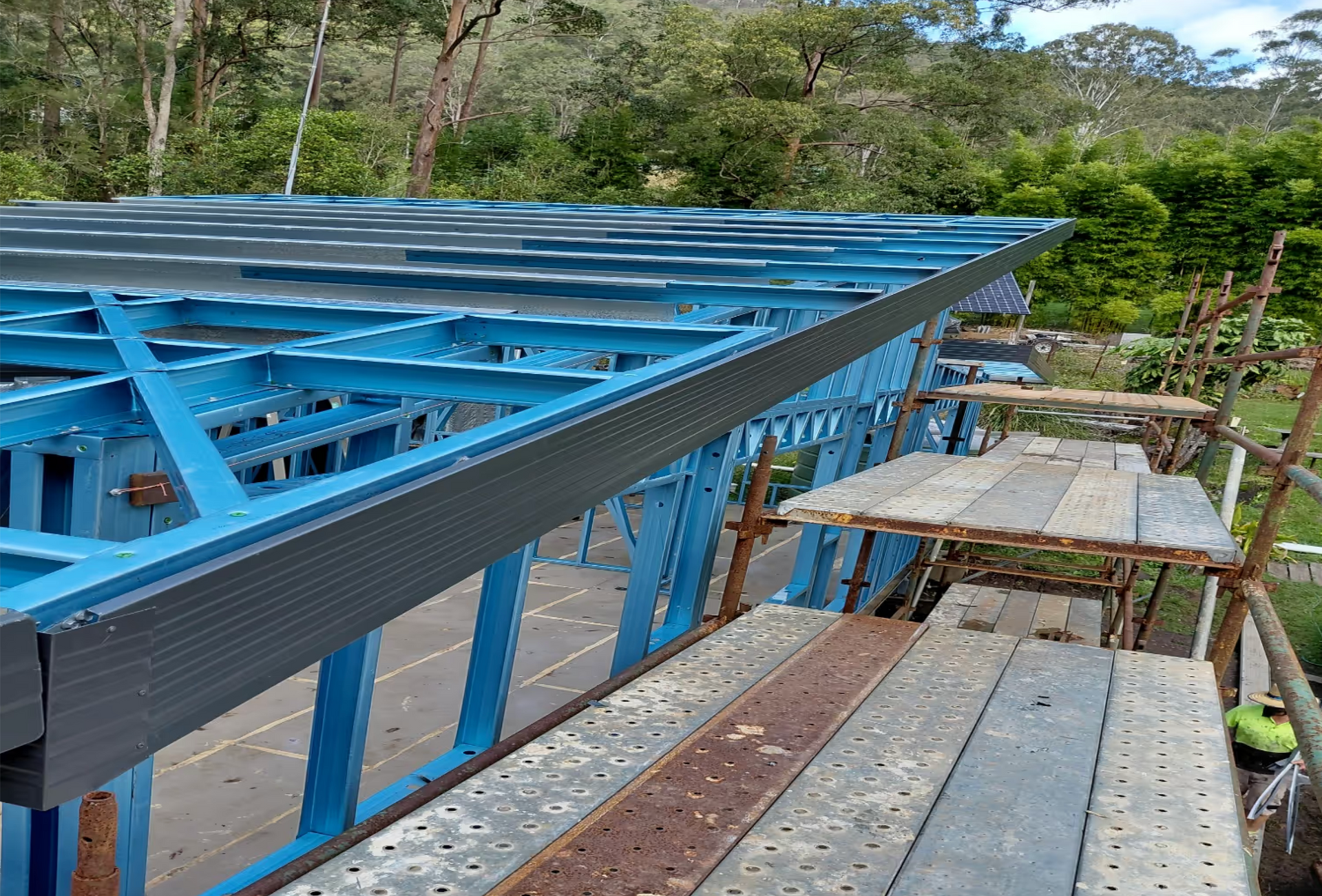 Blue steel roof framing beside a narrow wooden walkway in a wooded outdoor construction site