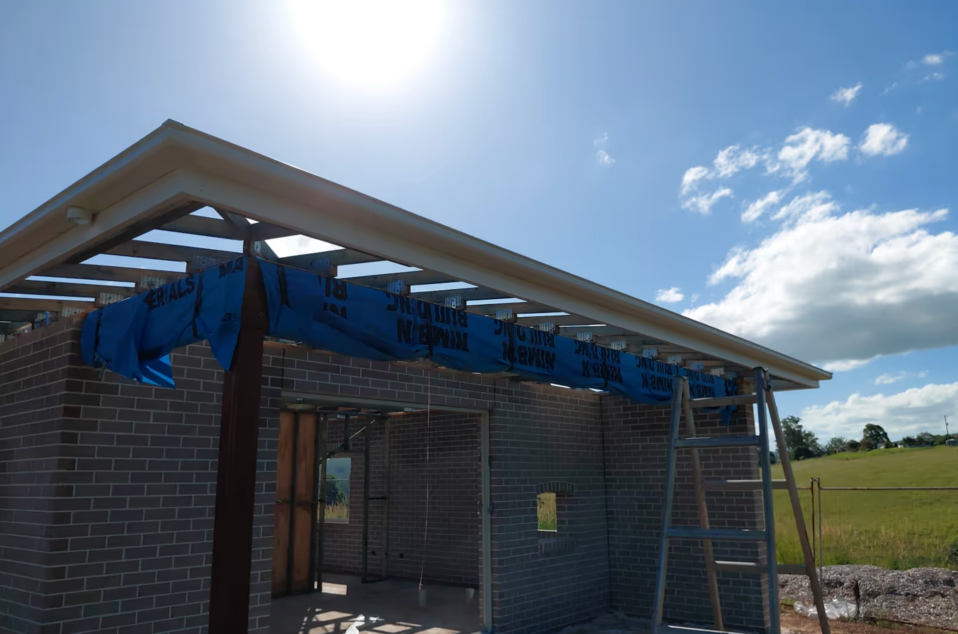 Partially built brick house with blue tarp and exposed roof under a bright sunny sky