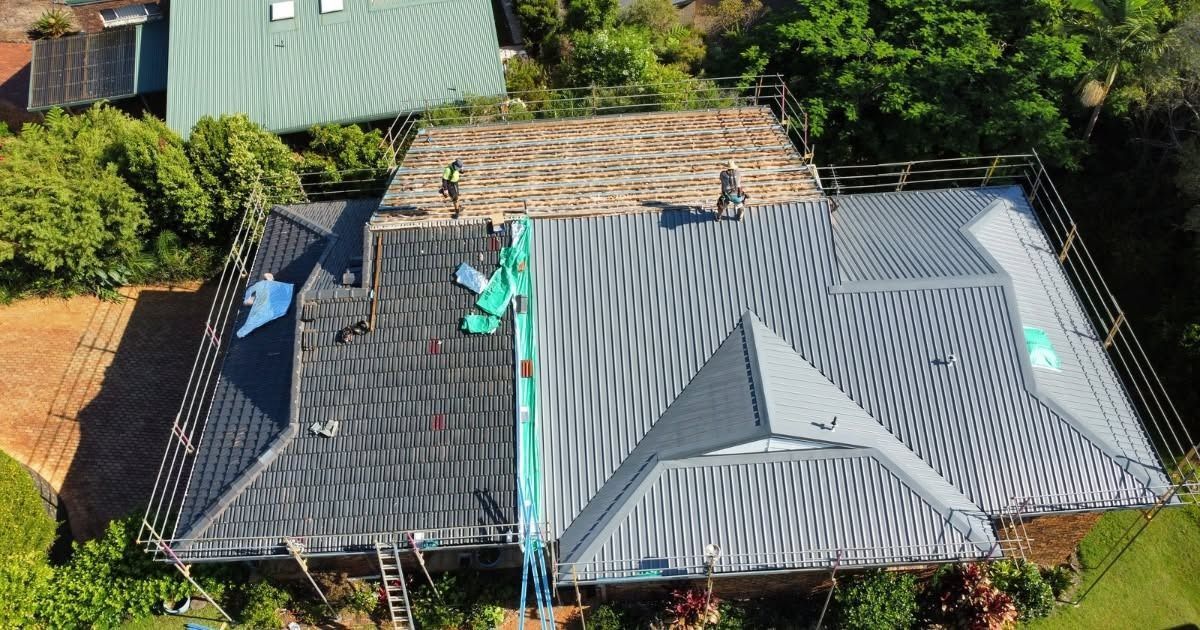 Aerial view of a house with a gray metal roof and solar panels, surrounded by green yard and trees
