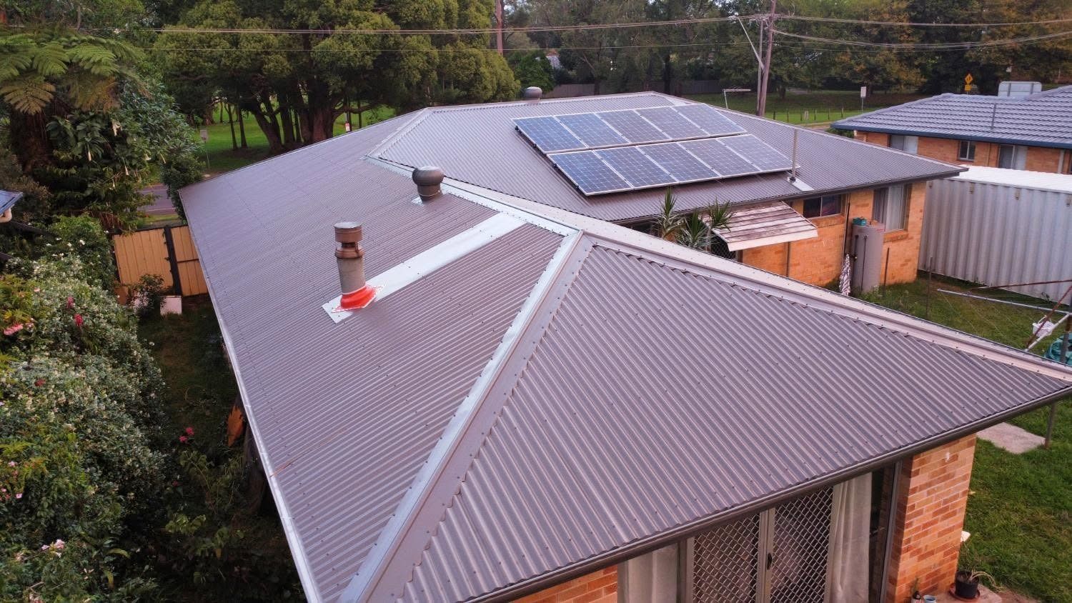 Aerial view of a gray metal-roofed house with solar panels and a chimney, surrounded by trees and neighboring homes.