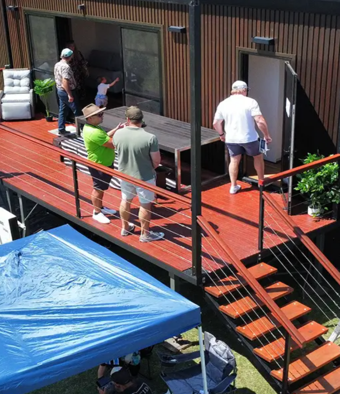 People on a wooden deck inspecting a building exterior near stairs and a blue canopy.