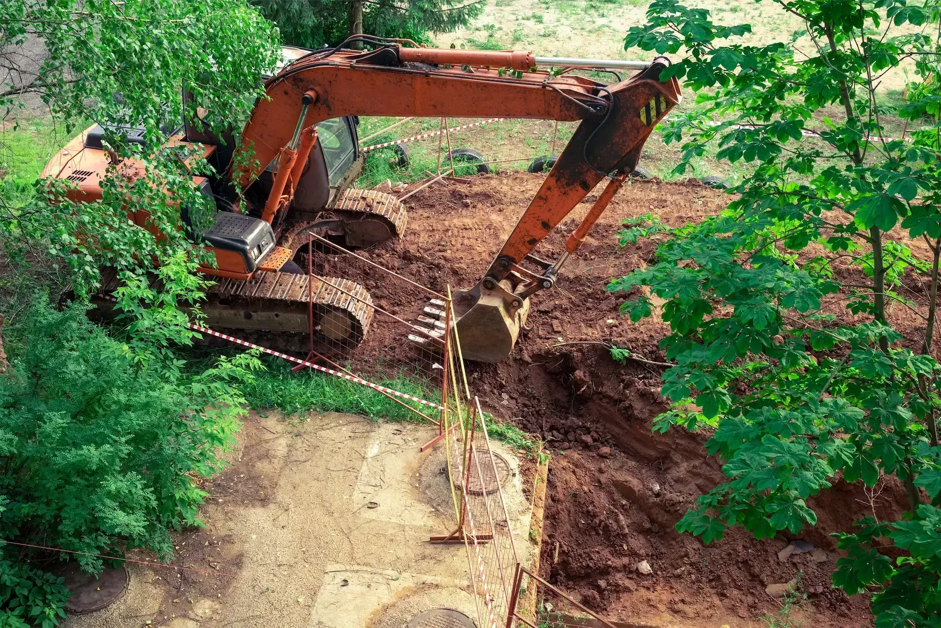 Excavator digging mud on a wooded construction site, with a worker nearby.