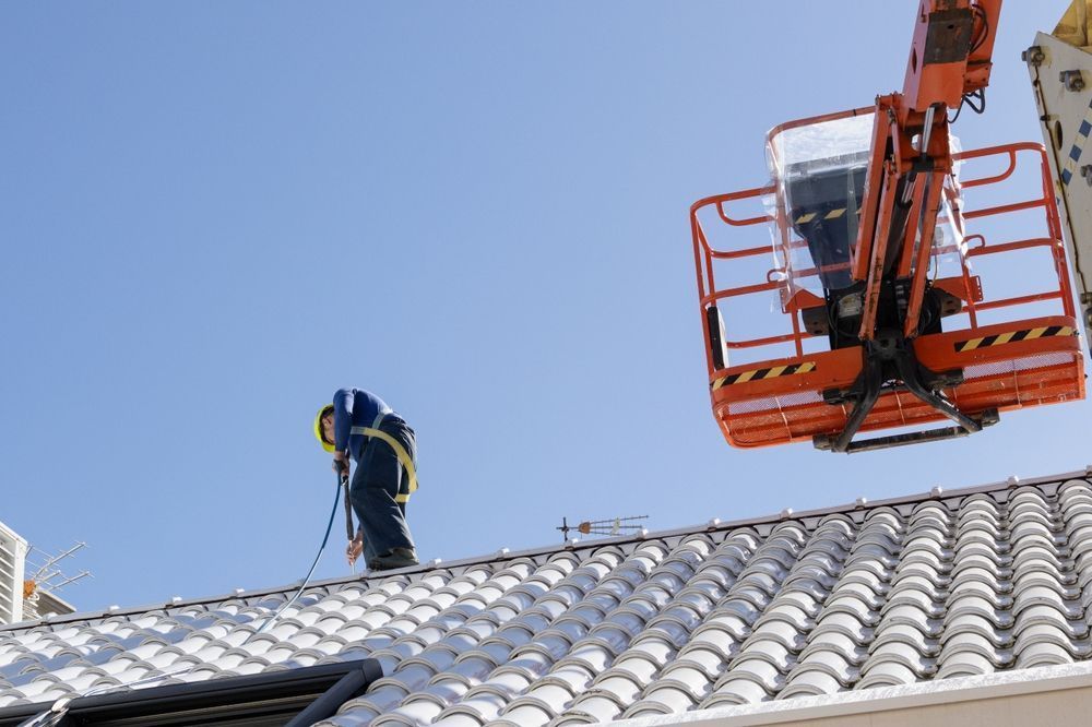 Worker in safety gear on a tiled roof beside an orange lift against a clear blue sky