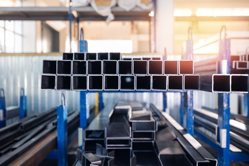 Stacked black metal tubes in a factory, suspended on blue racks with warm sunlight in the background