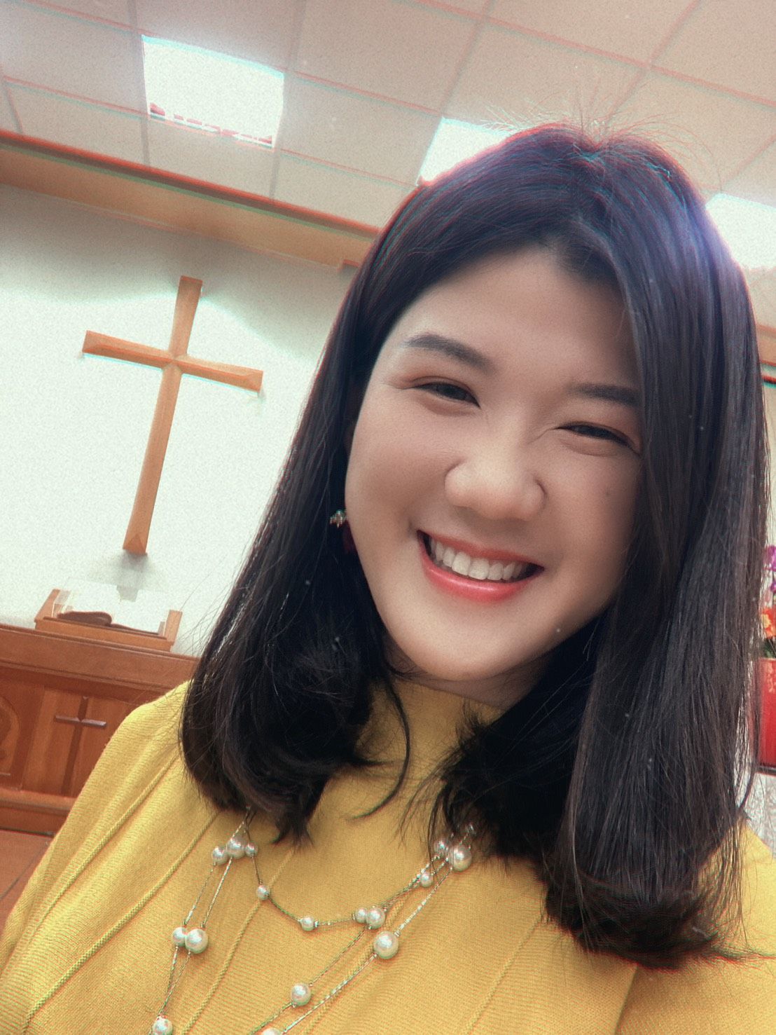 一位身穿黄色上衣、佩戴珍珠项链的微笑女子在房间里自拍,房间里的墙上挂着一个木制十字架。