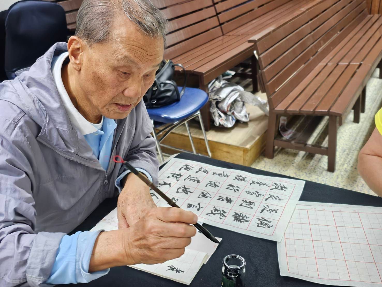 一个人正在桌边用毛笔和墨水在纸格上练习中国书法，桌旁有长凳。