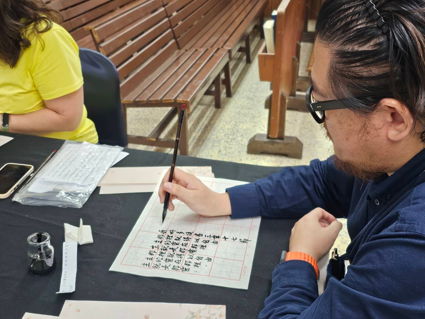 一名身穿蓝色衬衫的人在木凳旁的桌子上，用毛笔在方格纸上书写中国书法。