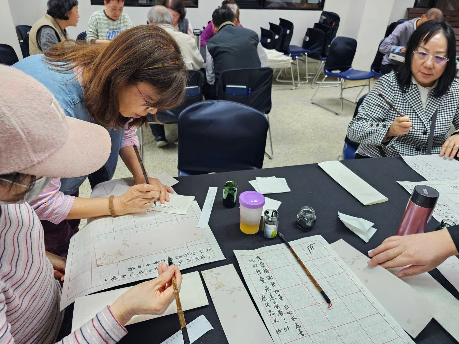 一群人在室内，坐在桌旁，在方格纸上练习传统书法。