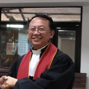 一位面带微笑的神职人员身穿黑色长袍,披着红色圣带,戴着白色神职领,在握手时做着手势。