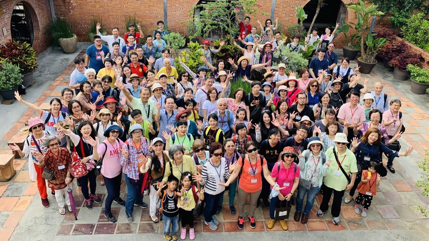 一大群不同背景的人们在砖墙环绕的户外庭院里摆姿势拍照,他们挥手微笑。