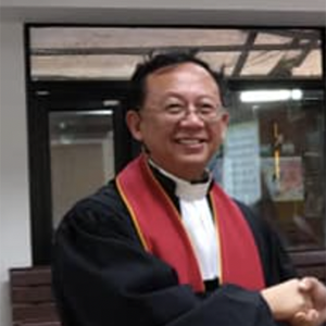 一位身穿黑色牧师长袍、披着红色圣带的人面带微笑,在室内与人握手。
