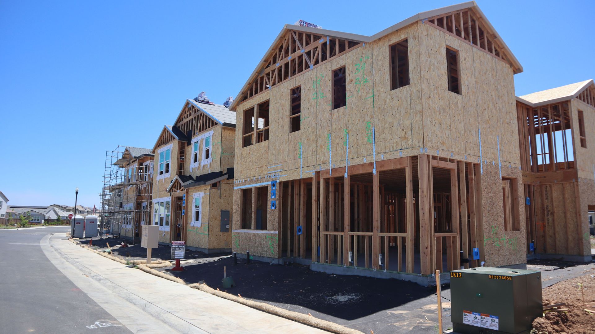 New suburban homes under construction on a sunny street, with exposed wood framing and blue sky.
