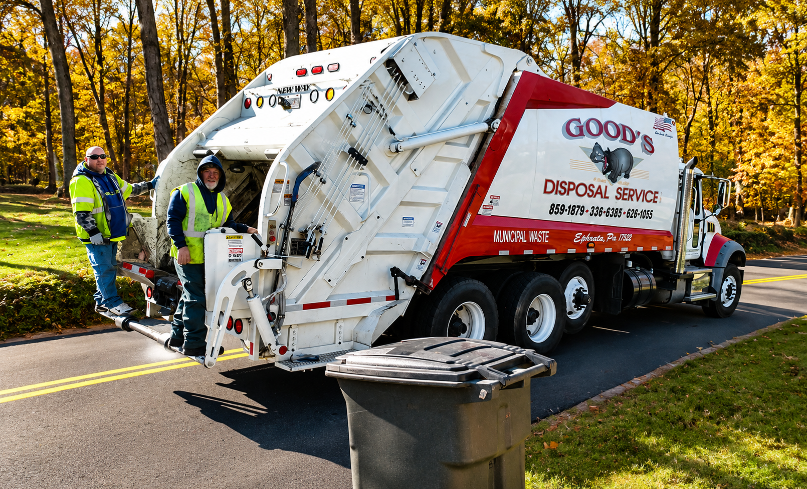 Garbage truck collecting trash on a suburban street with workers beside it and autumn trees in the background