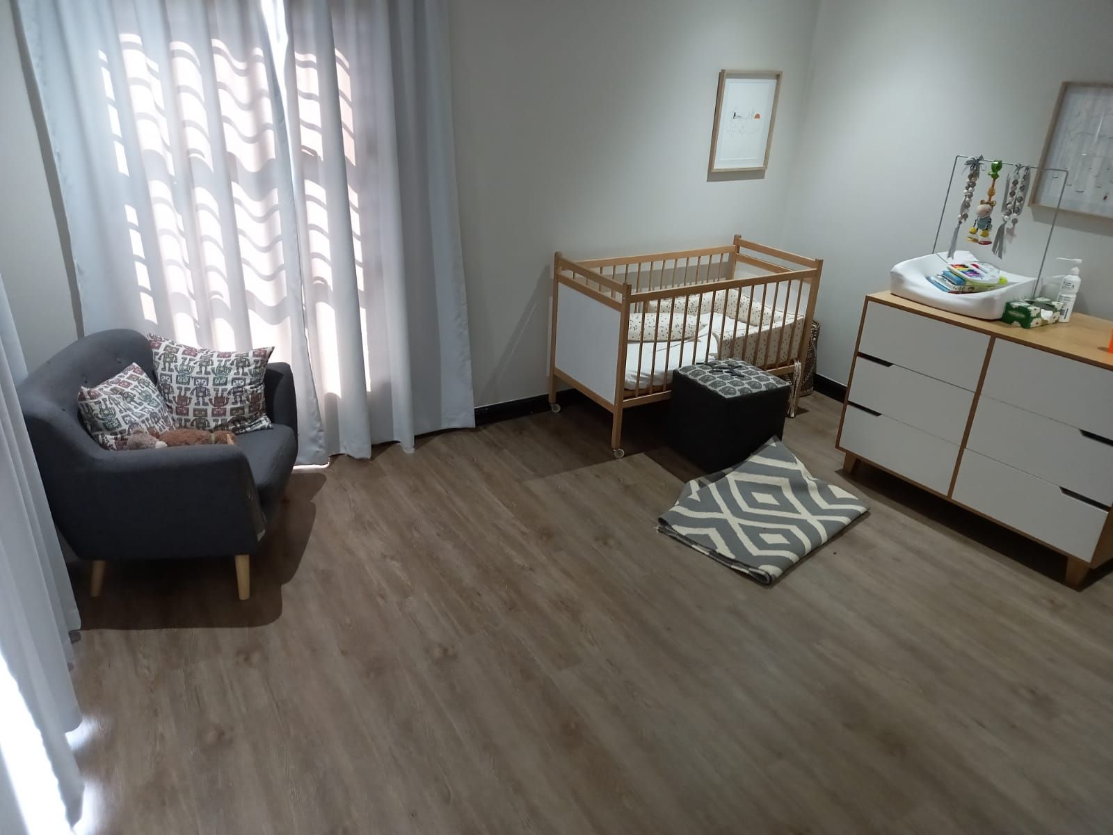 A baby room with a crib , dresser , chair and window.