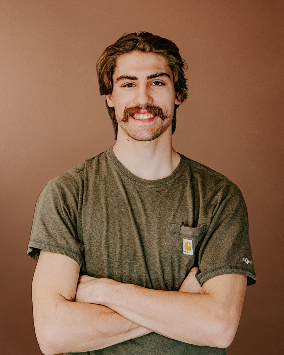 Smiling person with crossed arms wearing an olive T-shirt against a brown backdrop