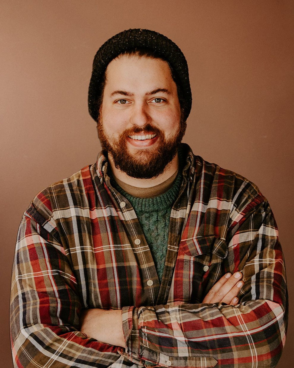 Smiling man with a beard and knit cap, arms crossed, wearing a plaid shirt against a plain brown background