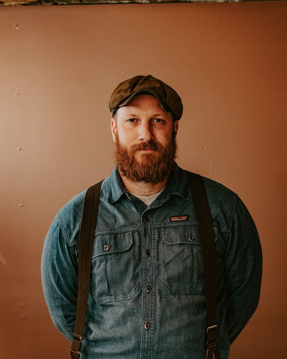 Portrait of a bearded man in a cap and denim overalls against a plain brown wall