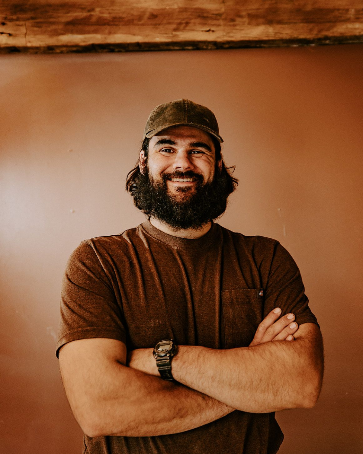Smiling bearded person in a cap, arms crossed, against a warm-toned wall.