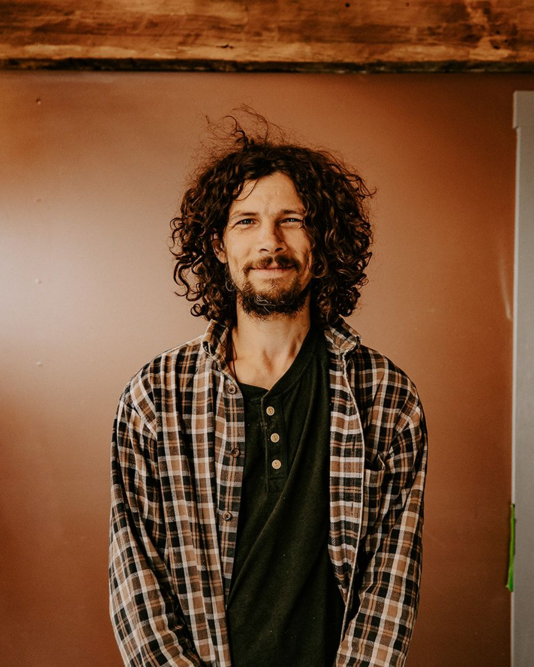 Smiling person with curly hair in a plaid shirt standing against a brown wall.