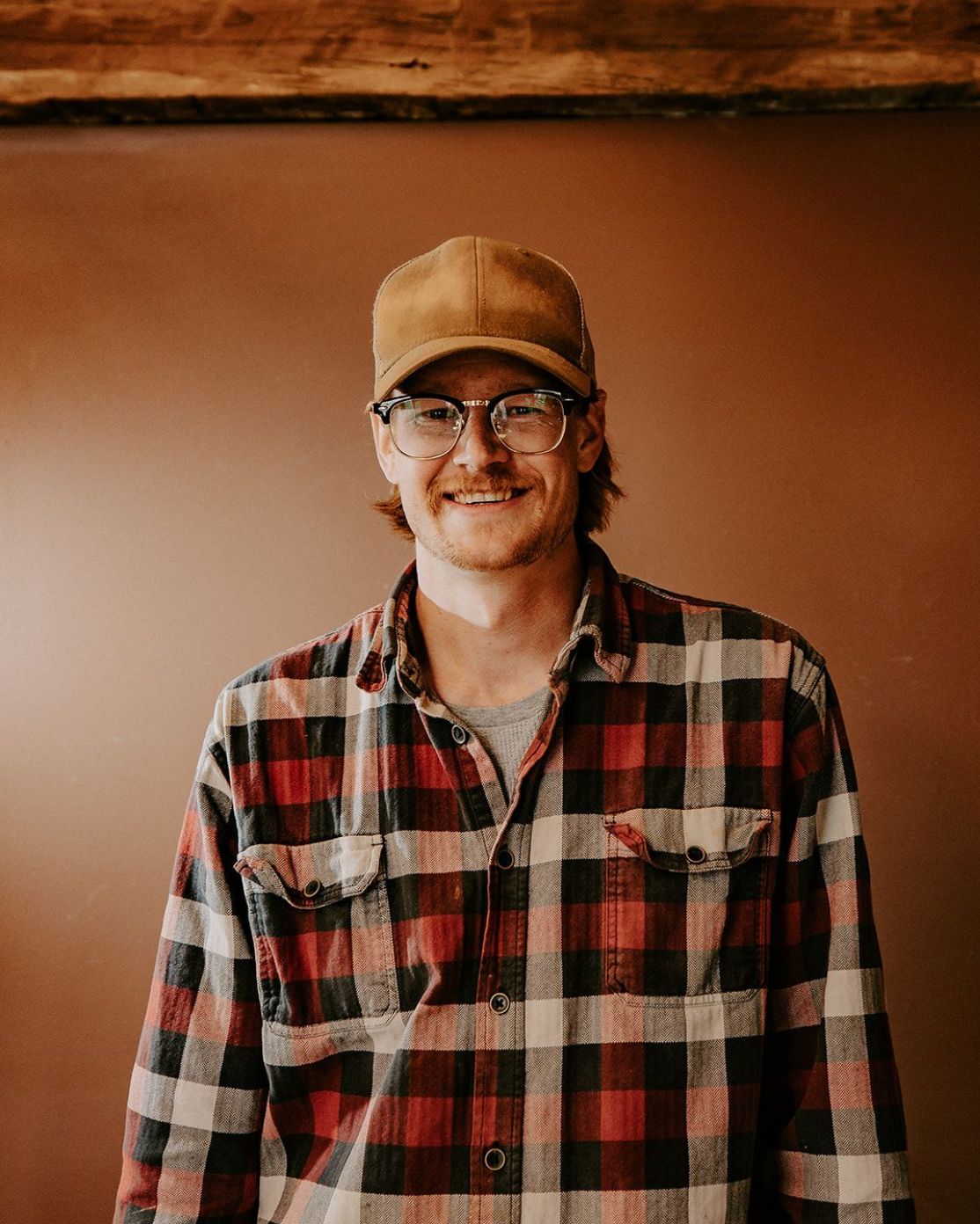 Smiling person in a red-and-black plaid shirt and tan cap standing indoors against a brown wall