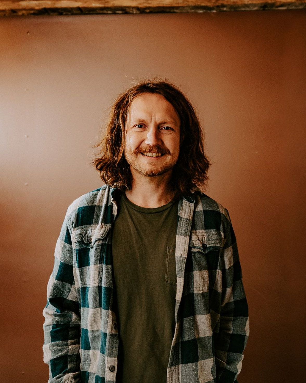Smiling person with shoulder-length hair wearing a plaid shirt, standing against a warm brown wall.