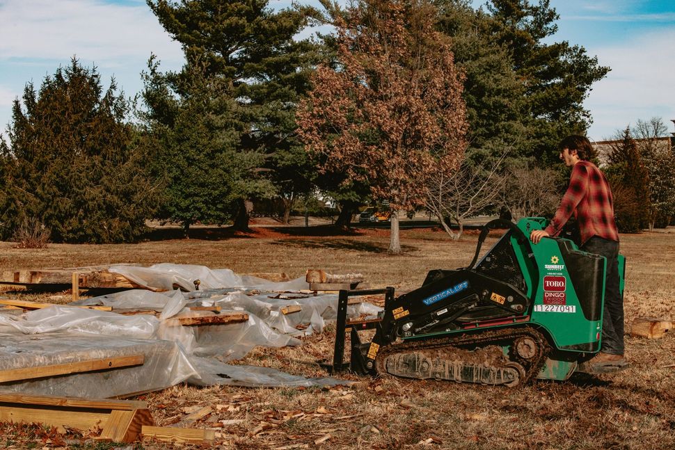 Person on a green skid steer moving wood chips in a yard with trees and covered materials nearby