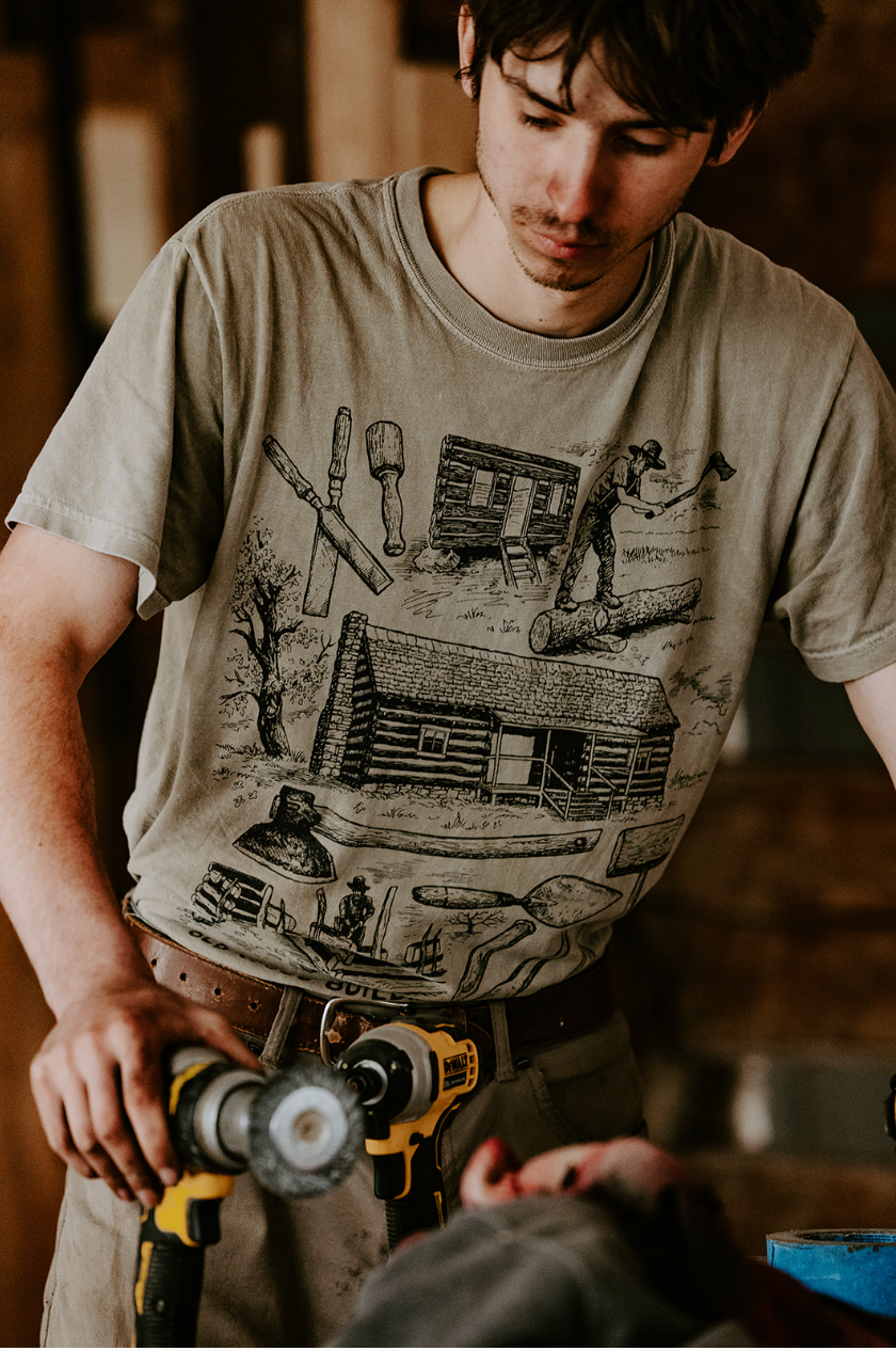 Person polishing a skateboard indoors, wearing a tan graphic T-shirt.