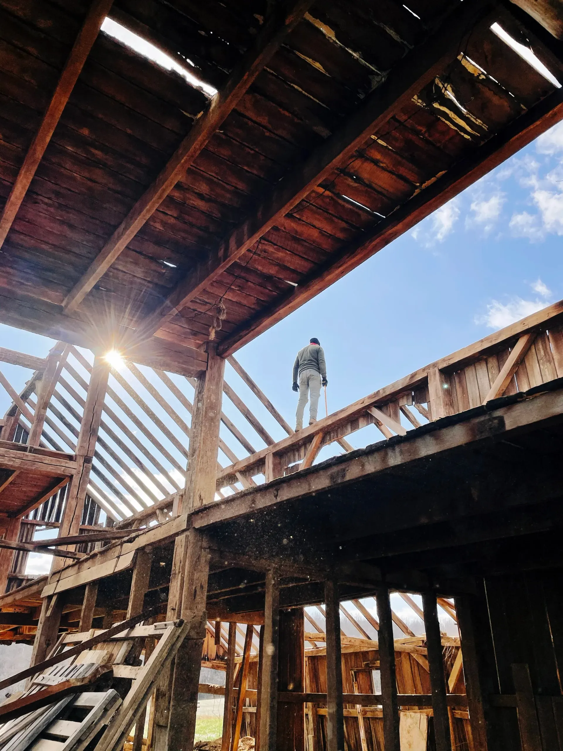 Person standing on a wooden beam in a sunlit, unfinished building frame with exposed rafters