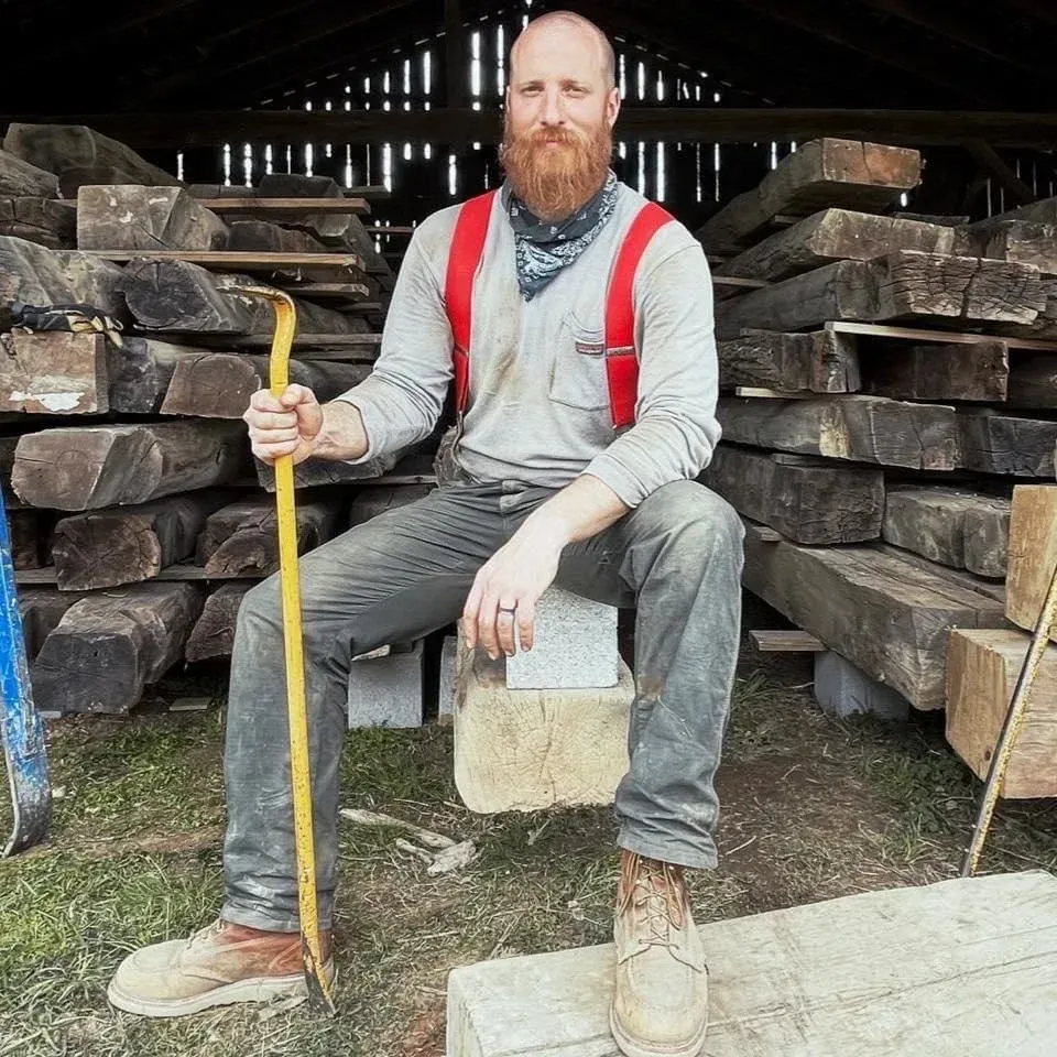 Person sitting on stacked timber in a workshop, holding a yellow tool and wearing red suspenders.