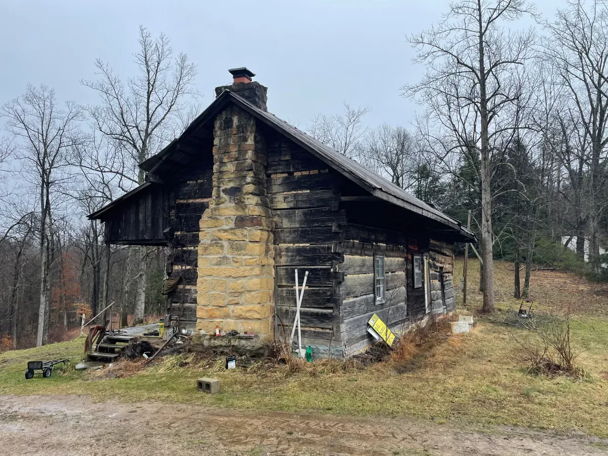 Burned, weathered cabin on a grassy hillside with bare trees in winter.